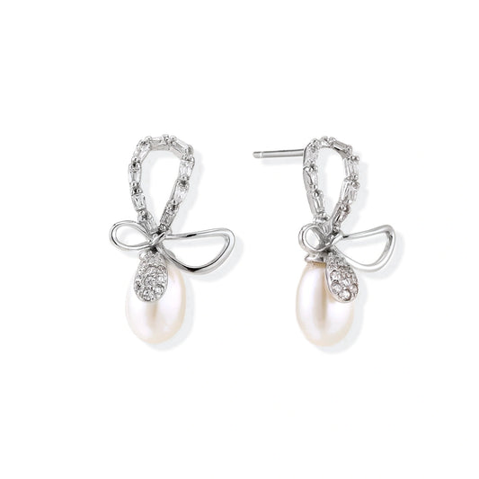 Silver bow-shaped stud earrings with baguette crystals and dangling white freshwater pearls on cream background