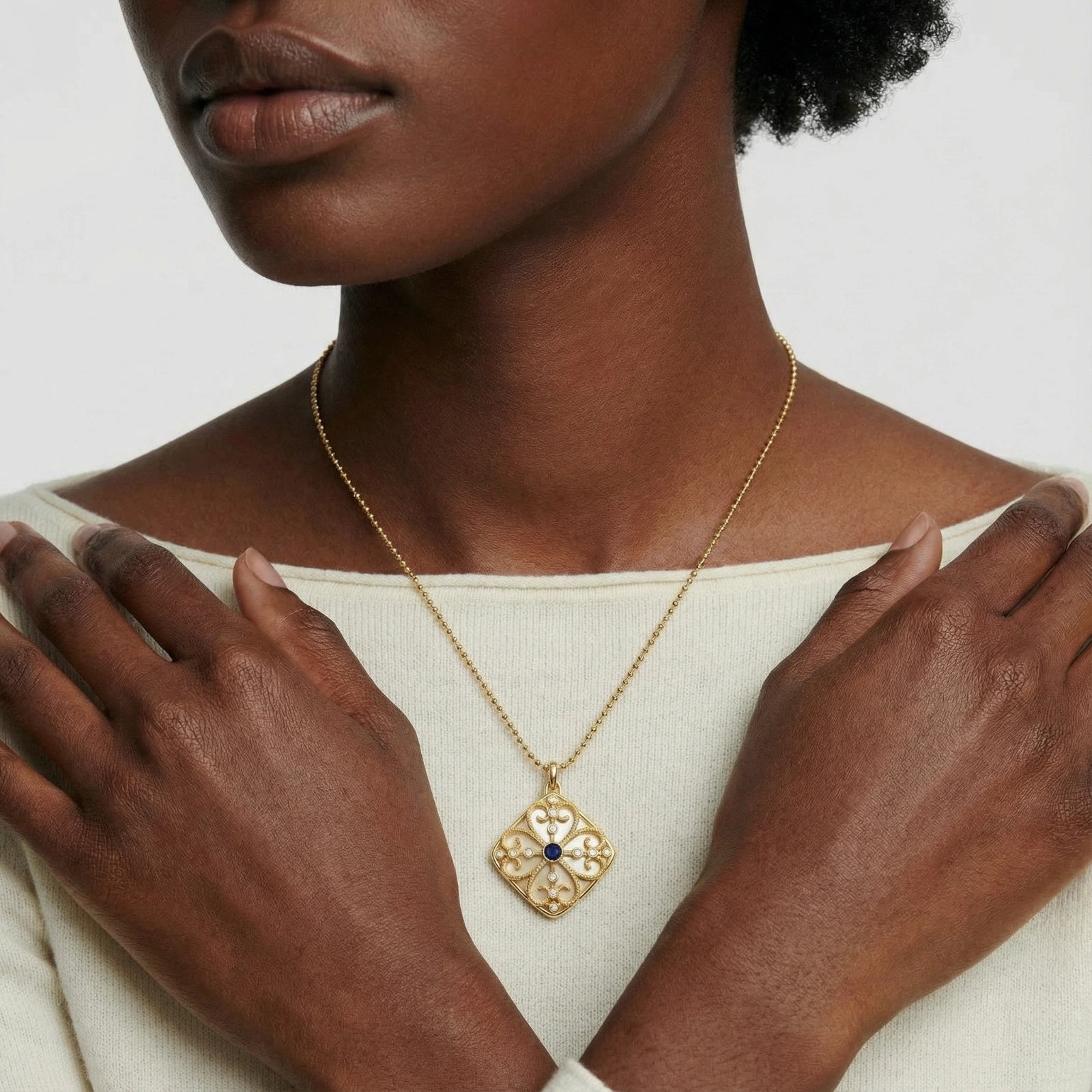 Woman wearing a 14K gold plated Wisdom Pendant necklace with intricate filigree design and a small dark gemstone in the center, styled with a cream-colored top.