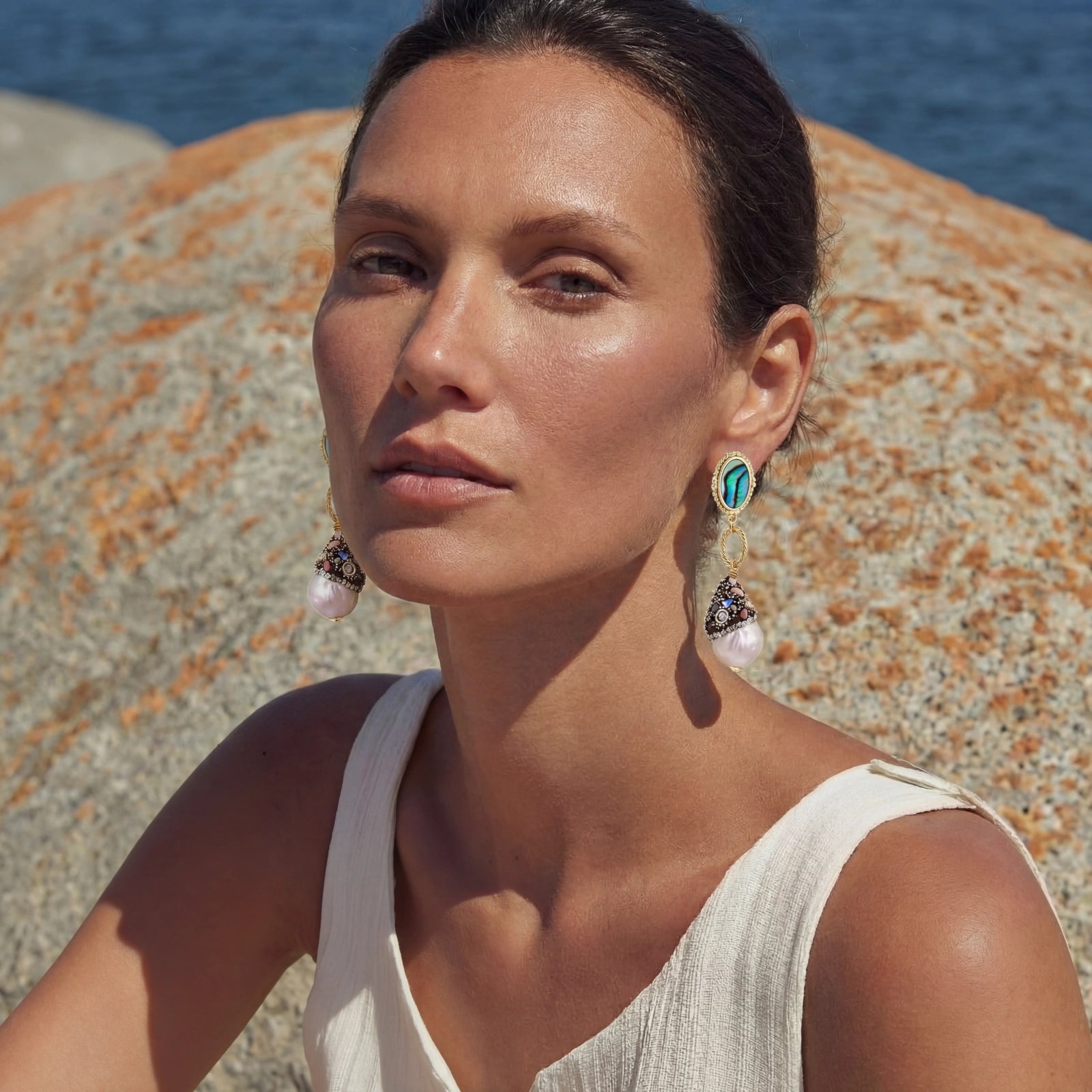 Woman wearing Baroque Pearl Statement Earrings with irregular pearls and gold plated accents, posing outdoors near rocky background and water.