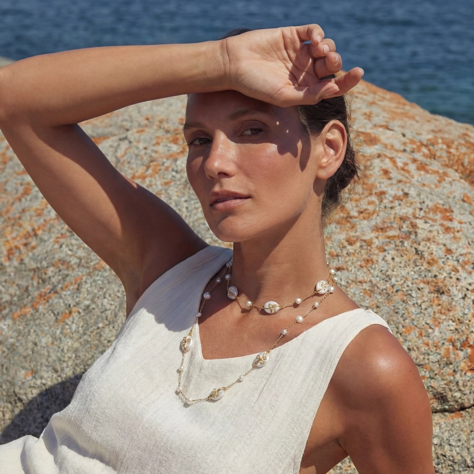Woman wearing a Baroque Pearl Garden Necklace featuring 14K gold plated accents and baroque pearls, styled with a sleeveless white top against a rocky seaside background.