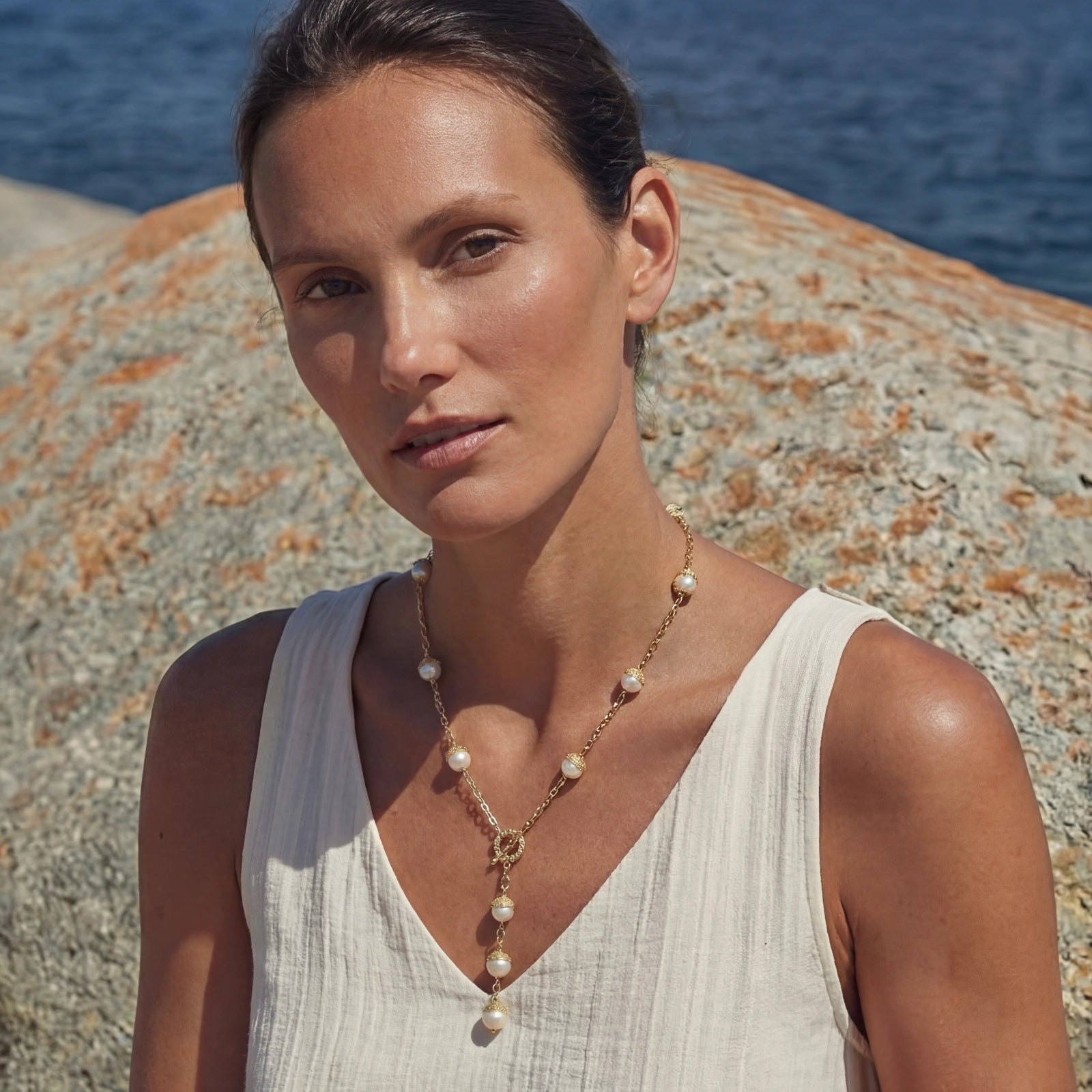 Woman wearing a Baroque Pearl Station Necklace featuring irregular baroque pearls set in 14K gold plated chain, styled with a white sleeveless top against a natural rocky seaside background.