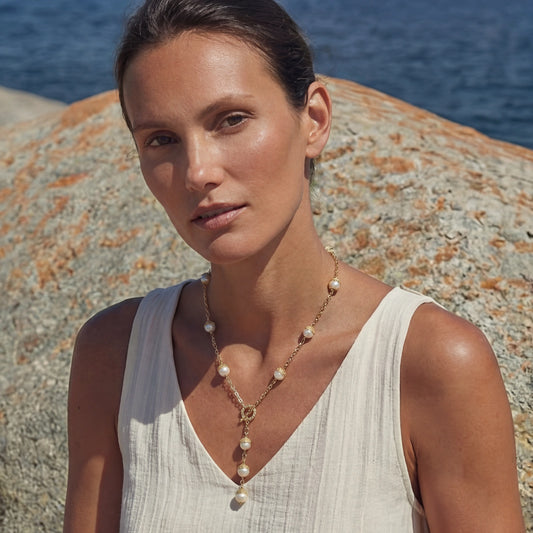 Woman wearing a Baroque Pearl Station Necklace featuring irregular baroque pearls set in 14K gold plated chain, styled with a white sleeveless top against a natural rocky seaside background.