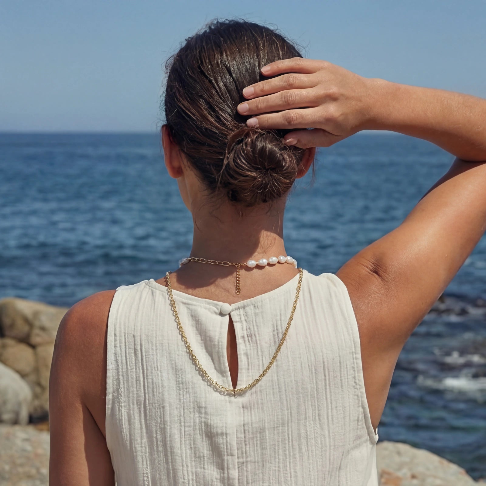 Woman wearing AlbaLuxe Golden Pearl Layers Necklace featuring 14K gold plated chain and baroque pearls, styled against a coastal backdrop with her hair in a bun.