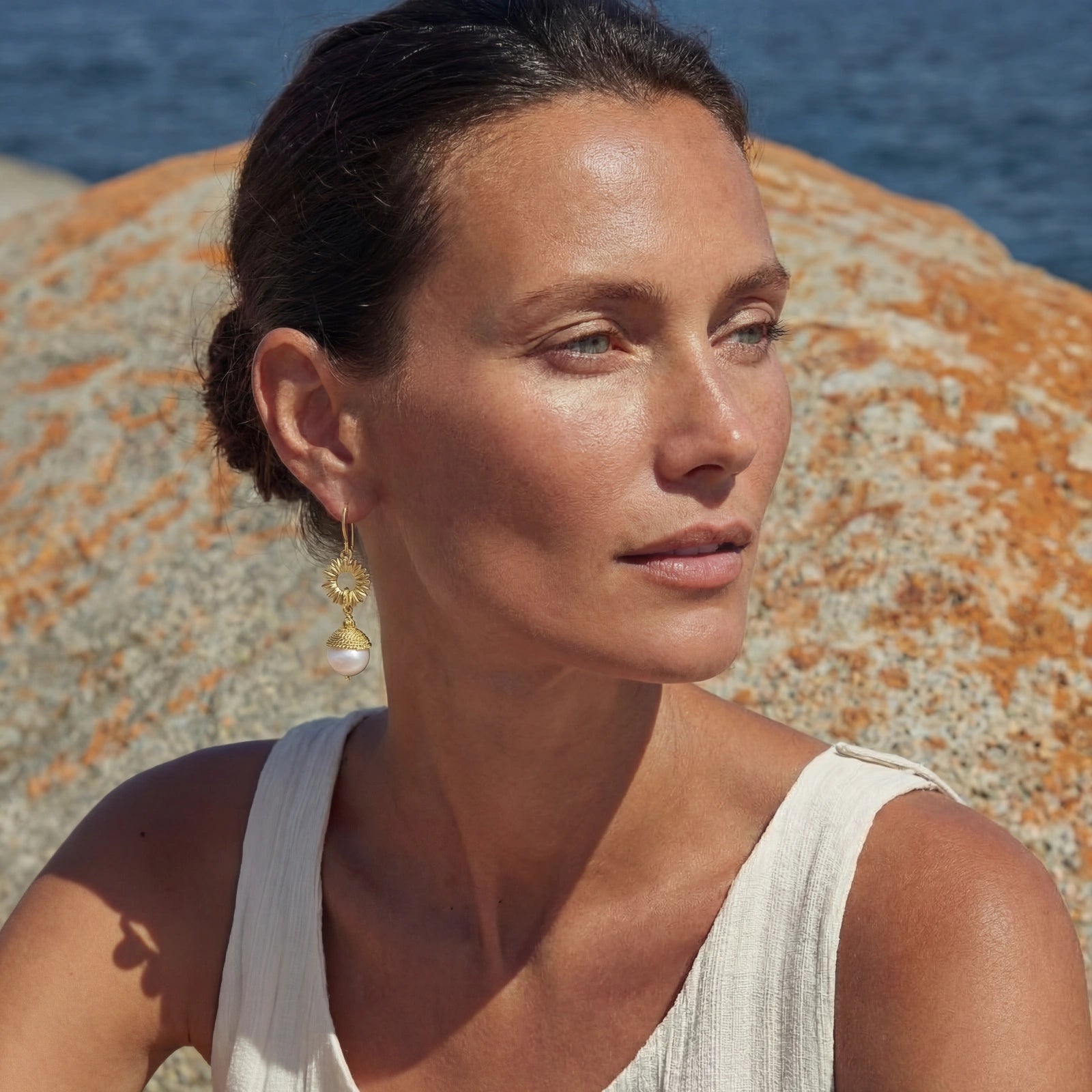 Woman wearing Sunburst Pearl Dangles featuring 14K gold plated sunburst design with a luminous pearl drop, styled outdoors by a rocky seaside background.