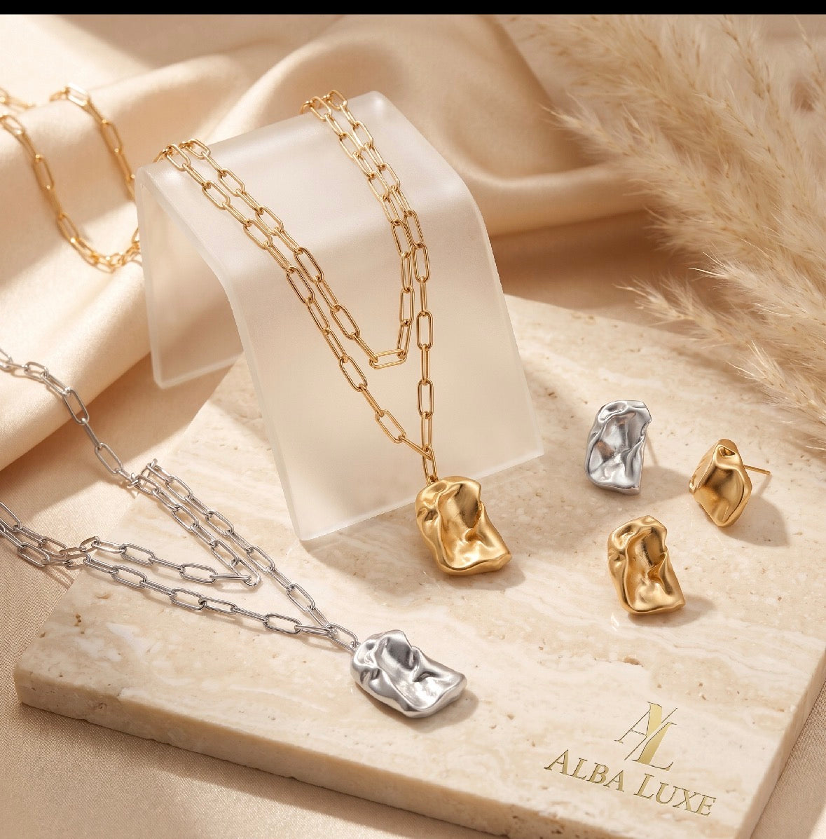 Two layered chain necklaces with irregular shaped pendants in gold and silver tones displayed on a clear stand, accompanied by matching gold and silver irregular textured stud earrings on a beige surface with soft fabric and dried pampas grass in the background.